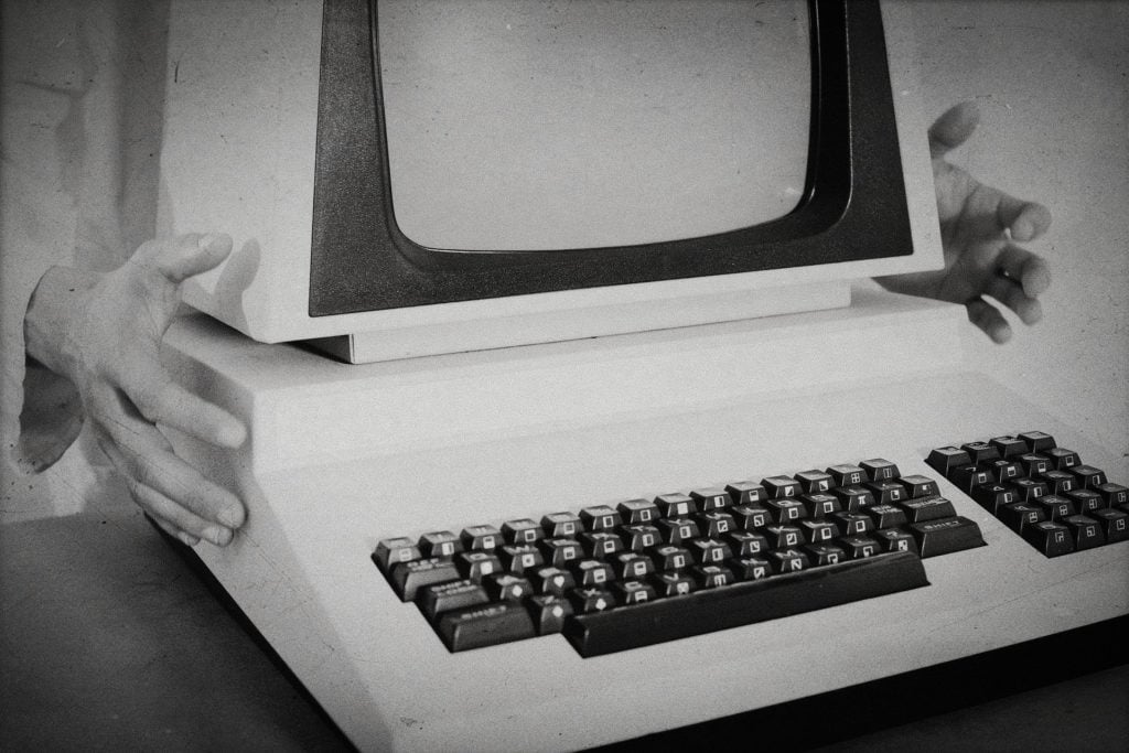 A black-and-white photo of a boxy, vintage computer with hands clutching it on either side from behind.