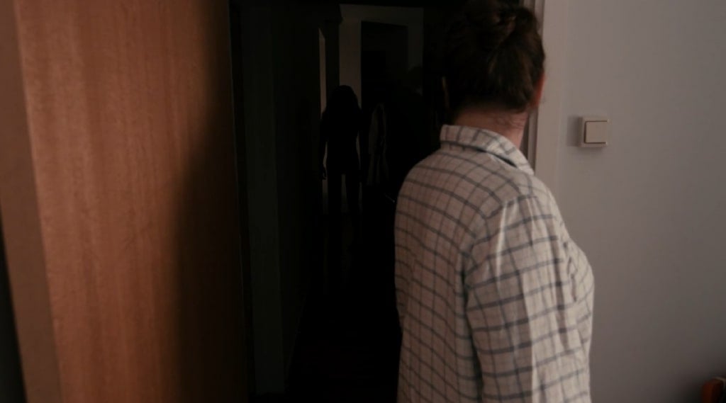 A screenshot from the short film "Lights Out" showing a female-presenting person looking down a dark hallway, where a figure lurks at the other end