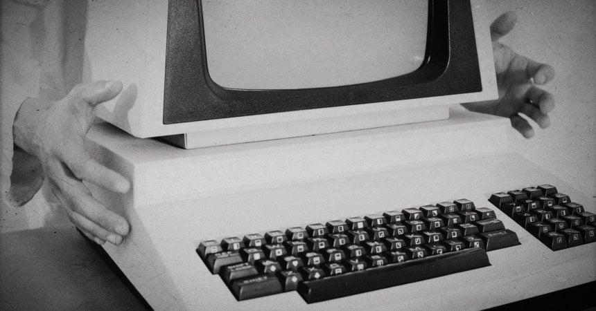 A black-and-white photo of a boxy, vintage computer with hands clutching it on either side from behind.