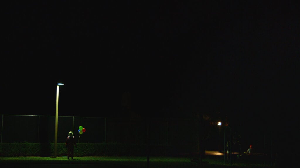 A screenshot of Wrinkles standing alone outside at night holding balloons from the Wrinkles the Clown documentary. Photo courtesy of Magnet Releasing.