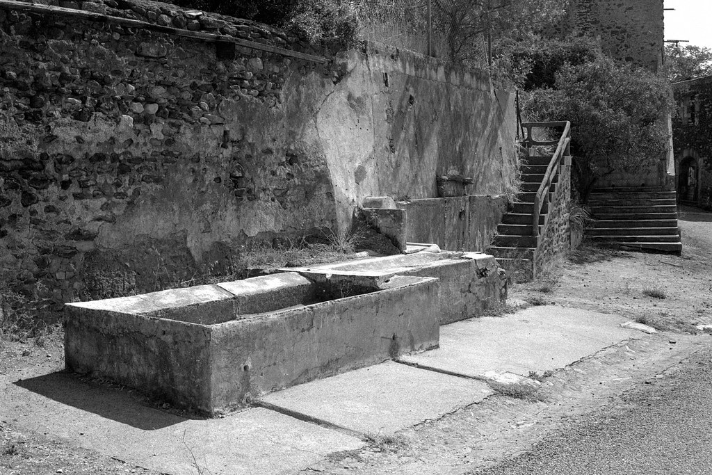 A black-and-white image of the exterior wall of a stone building with stone stairs, along with a stone or concrete cistern or similar out front.