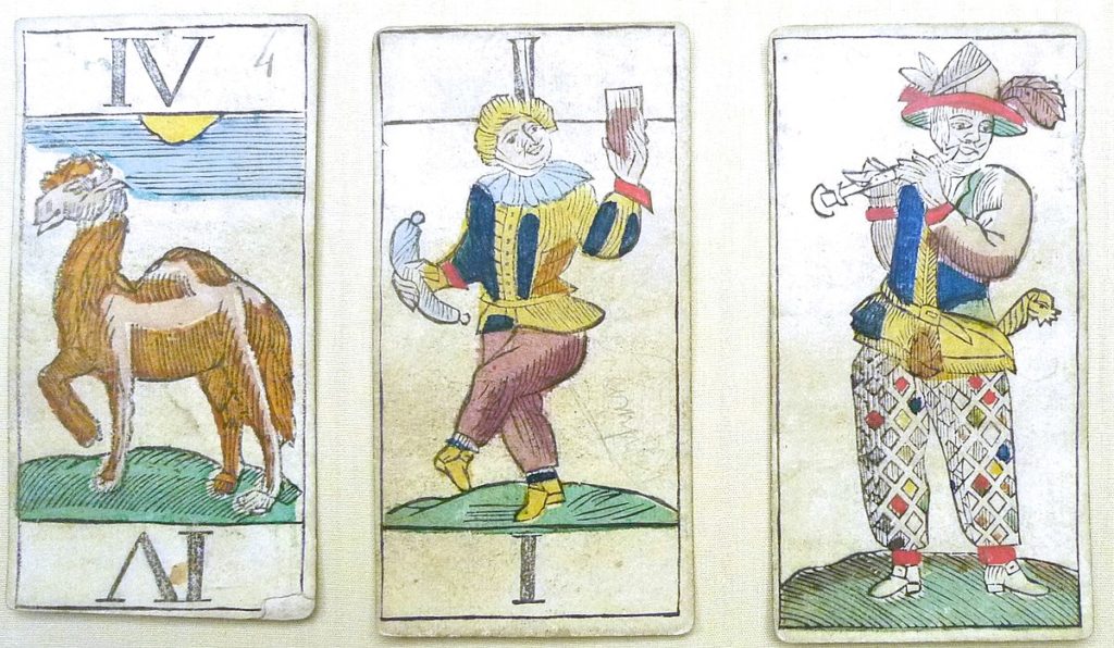 Three cards from a 16th century Belgian animal tarot deck: From left to write, a card with a camel numbered four, a card with a person holding a sausage and cup of wine numbered one, and a person in motley playing a pipe that isn't numbered.