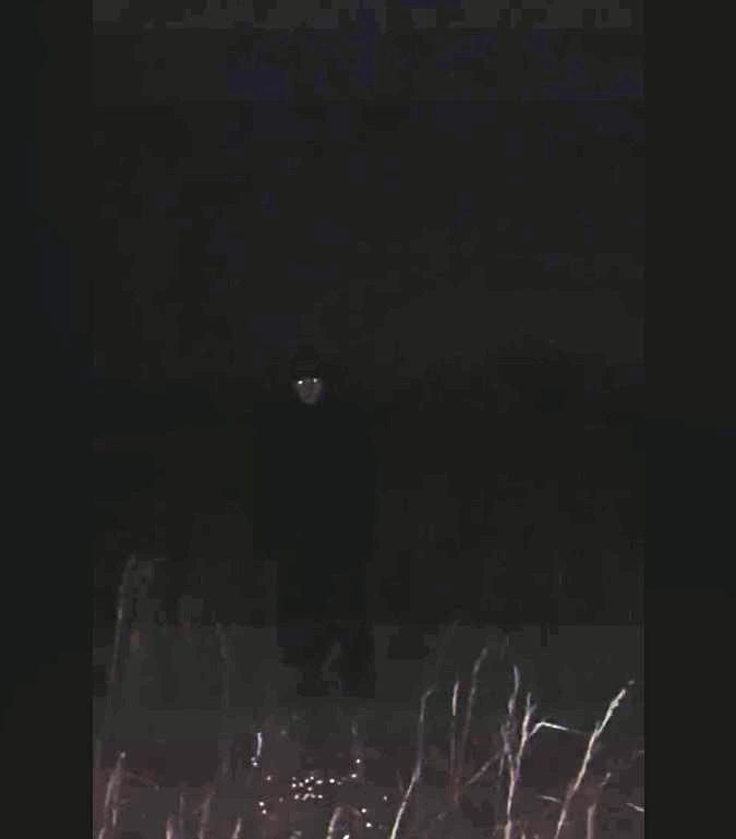 A brightened up version of the previous image. As before, it's a dark field at night with a humanoid figure standing at distance. The figure has glowing eyes.