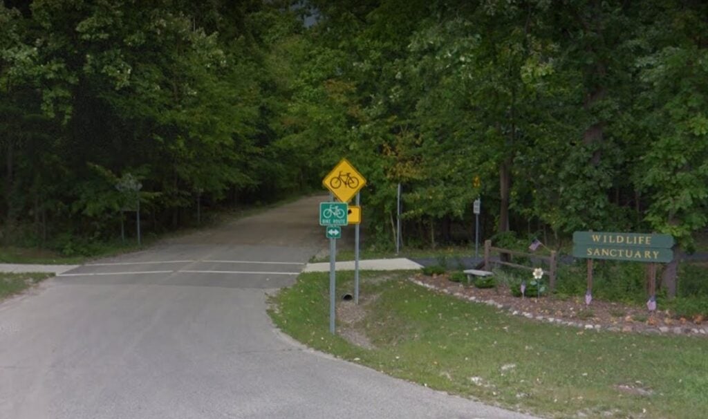 A road leading into a wooded area that's part of the GRosse Ile Wildlife Sanctuary. To the right, a green, wooden sign reading "Wildlife Sanctuary."