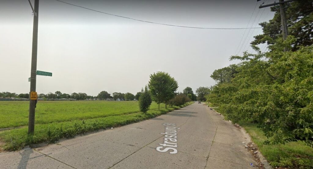 A stretch of road with grass on one side and leafy trees on the other. A street sign on a pole to the left reads "Strasburg."