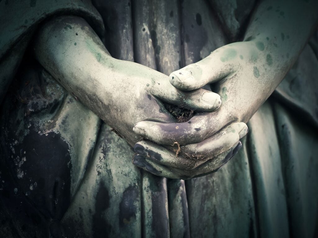 The hands of a statue in a cemetery, weathered and clasped together.