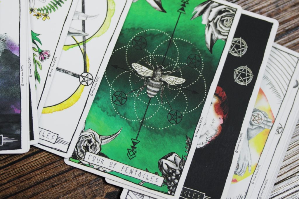A modern tarot deck, fanned across a wooden surface. The card visible on the top is green and decorated with a drawing of a bee. It represents the Four of Pentacles.