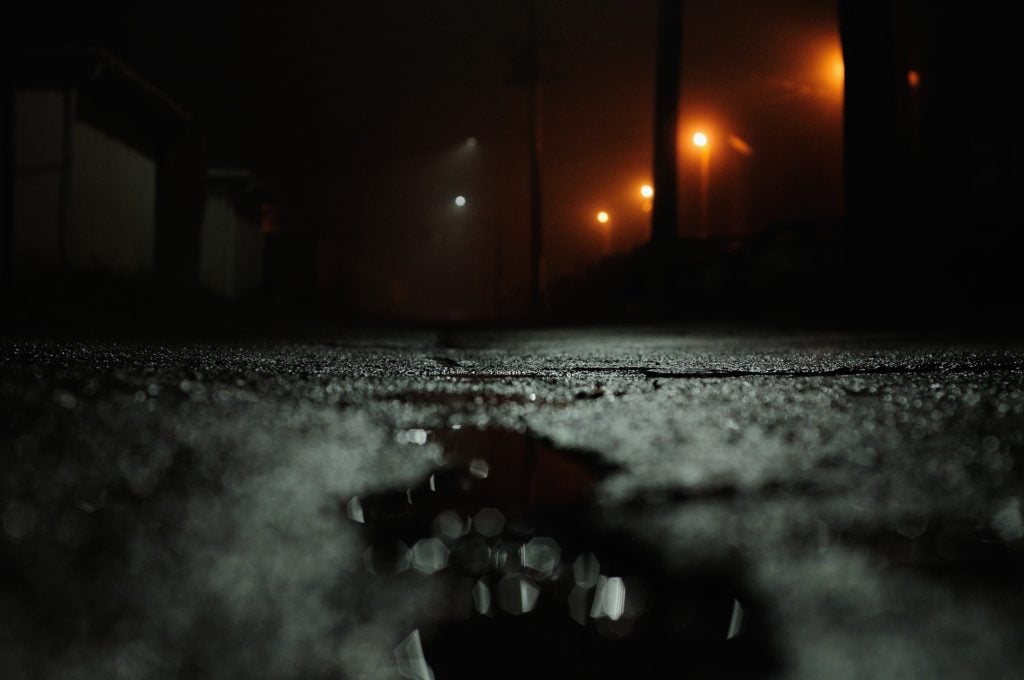 A dark, rainy street, with glowing streetlights in the distance.