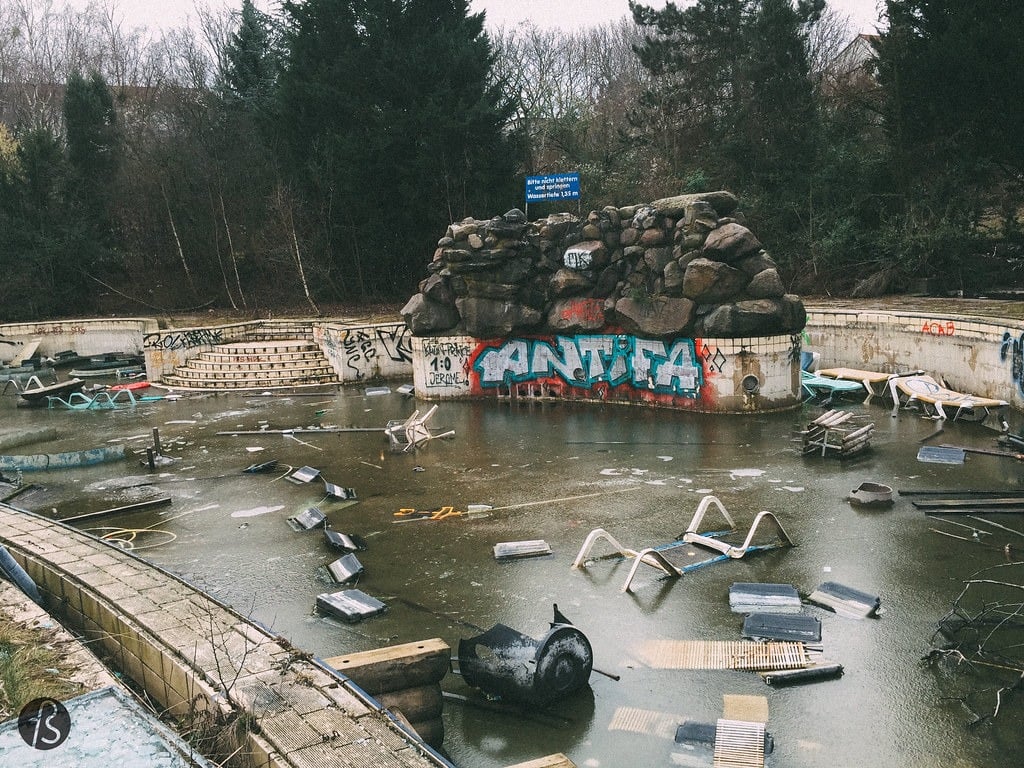 A large, abandoned outdoor pool at Blub water park in Berlin in 2015. The pool is empty of water, but full of trash. Fake rocks with graffiti are in the center.