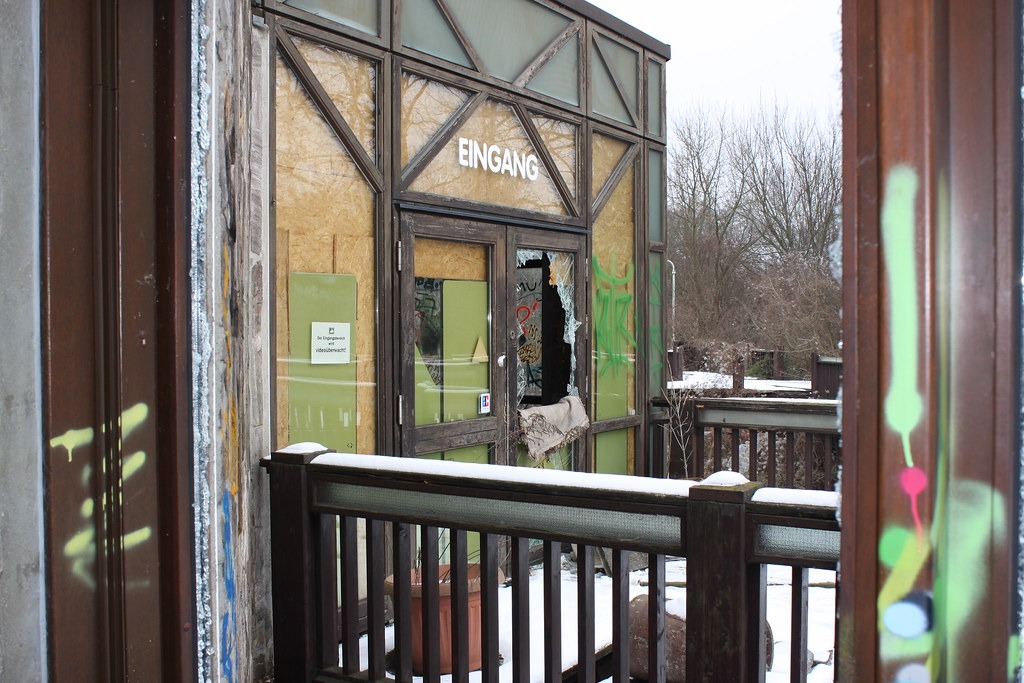 The remains of a wood and glass door with the word "EINGANG" written above it, viewed from the outside, at the abandoned Blub water park in Berlin, 2015.