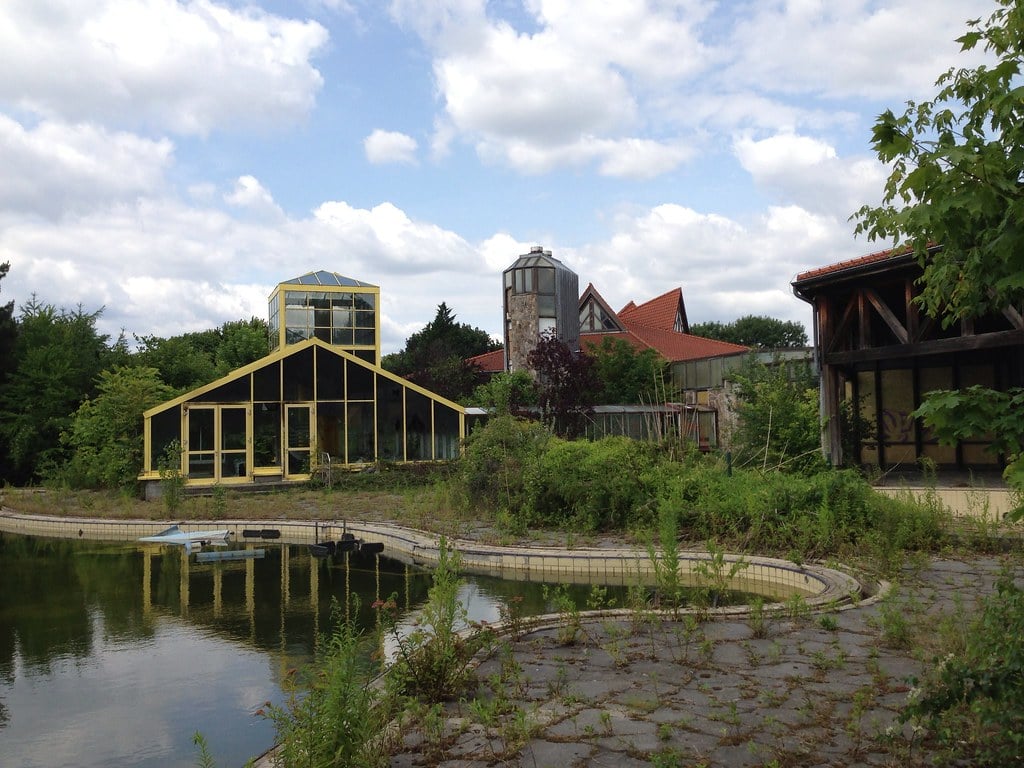 An overgrown outdoor pool with a peaked glass building, a tower, and two wooden buildings with red roofs in the distance at the abandoned Blub water park in Berlin, 2013.