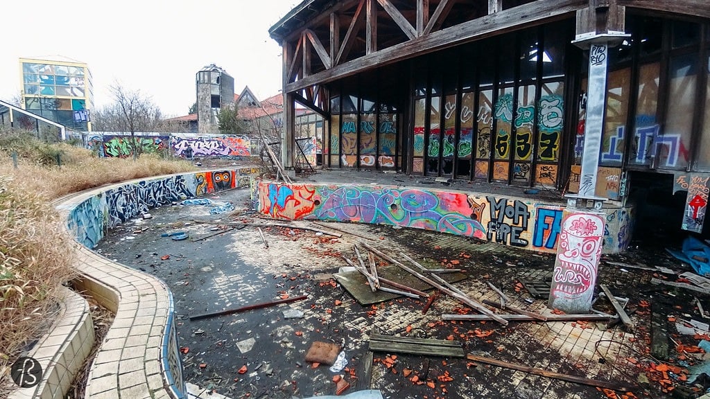 The same overgrown, empty, outdoor pool with a glass and stone tower in the distance at the Blub abandoned water park in Berlin, 2015. This time, it's covered in graffiti.