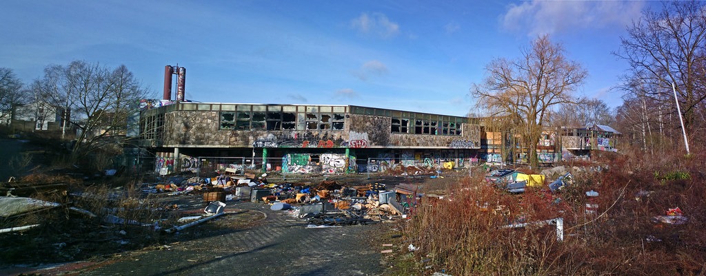 A panoramic shot of the exterior of the abandoned Blub water park in Berlin, 2017. It is a low building with stonework on the outside. The windows are all punched out. There is lots of litter and trash on the ground.