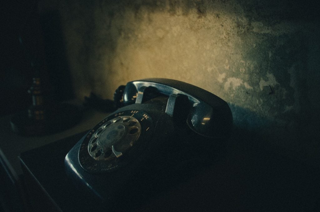 A vintage, black rotary telephone sitting on a dark surface in front of a grotty wall.