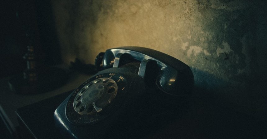 A vintage, black rotary telephone sitting on a dark surface in front of a grotty wall.