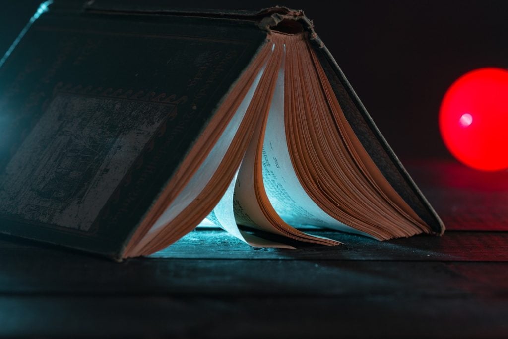 An old, hardcover book open and face down on a wooden surface with a light visible beneath its pages.