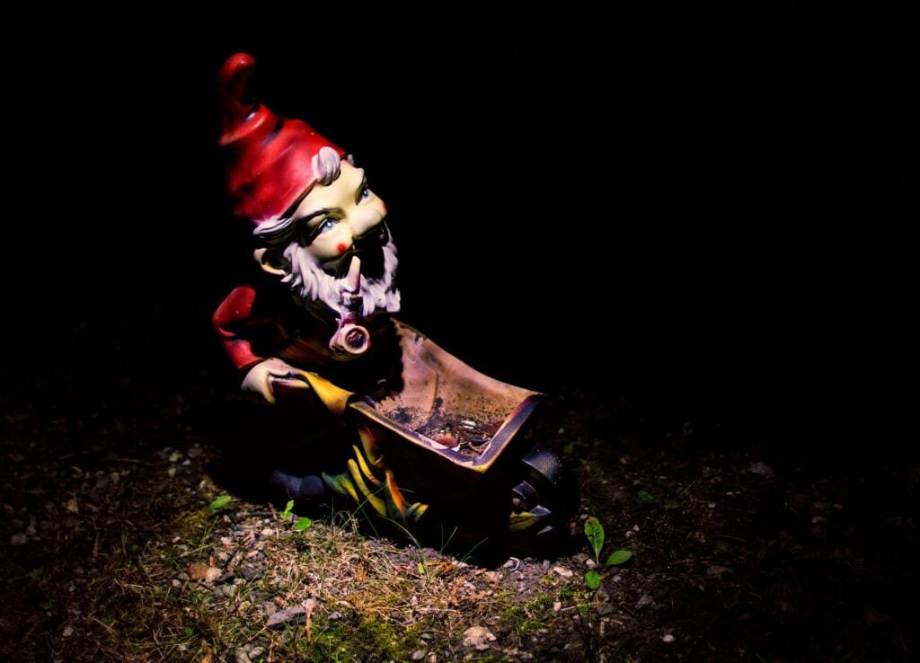A red-clad garden gnome sitting on the ground in the dark. It would be cute if it weren't so creepy.
