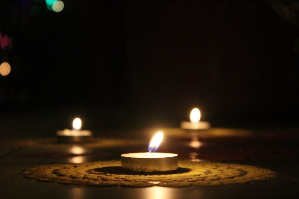 Three tea lights - one in the foreground, two in the background - sitting on lace doilies and burning in a dark room.