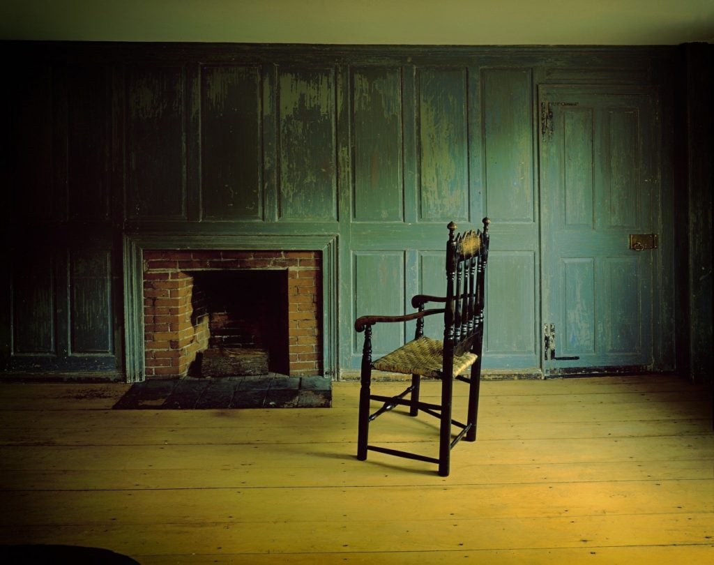 An old fireplace set in a green wall in a run-down, abandoned room. There's one wooden chair on the floor, but the room is otherwise empty and forlorn.