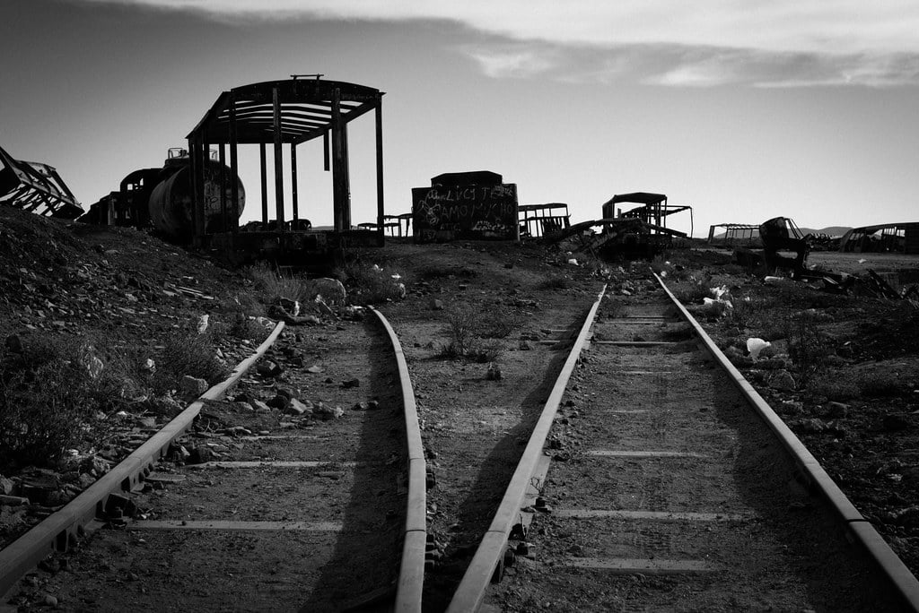 A black-and-white photo of two forking train tracks, abandoned in the desert. The frames of old train cars sit in the distance.