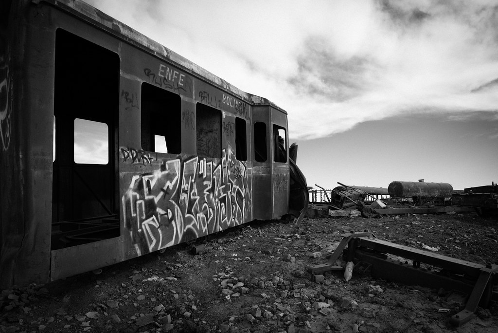 A black-and-white photo of an abandoned train car, covered with graffiti and with no wheels, sitting in the desert.
