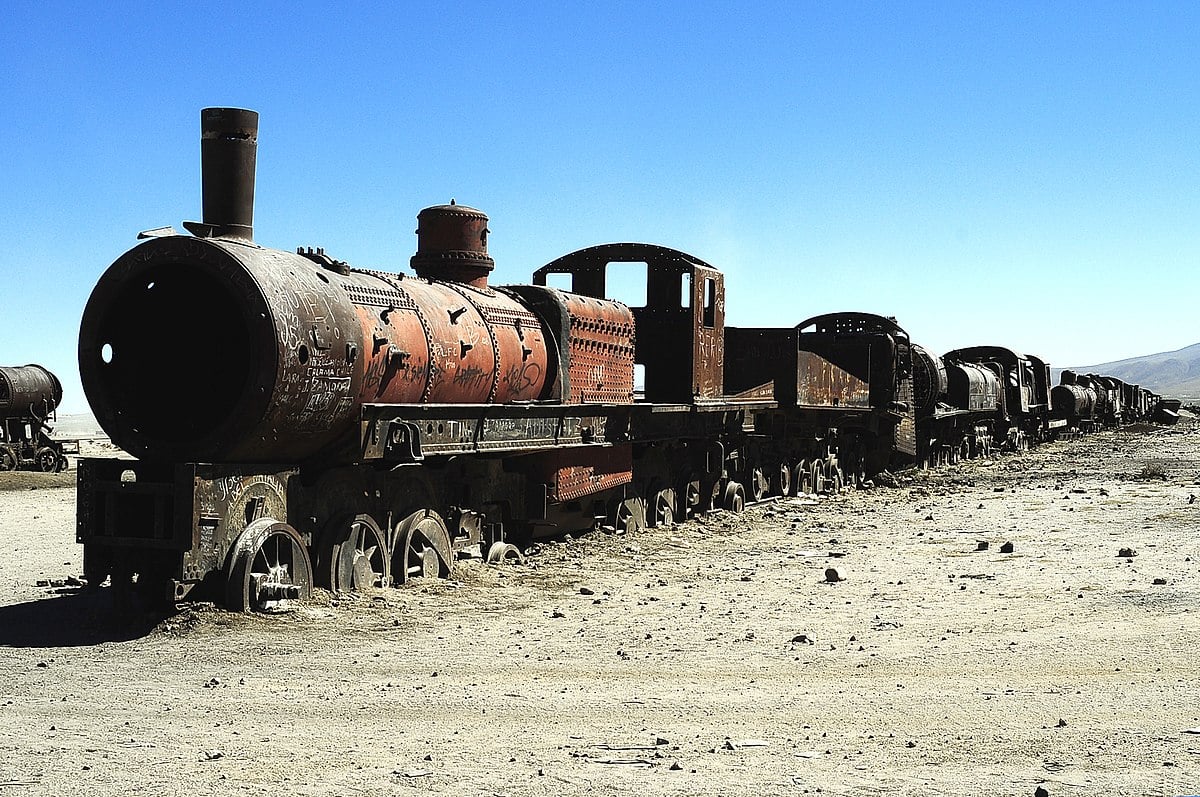 Abandoned train cars and an engine against a blue sky in the desert.
