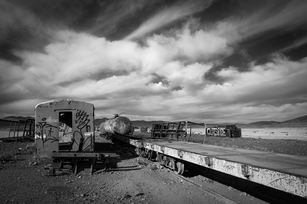 A black-and-white photo of an abandoned flat bed train car in the desert.