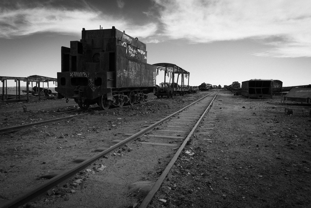 A black-and-white photo of an abandoned train car and tracks.