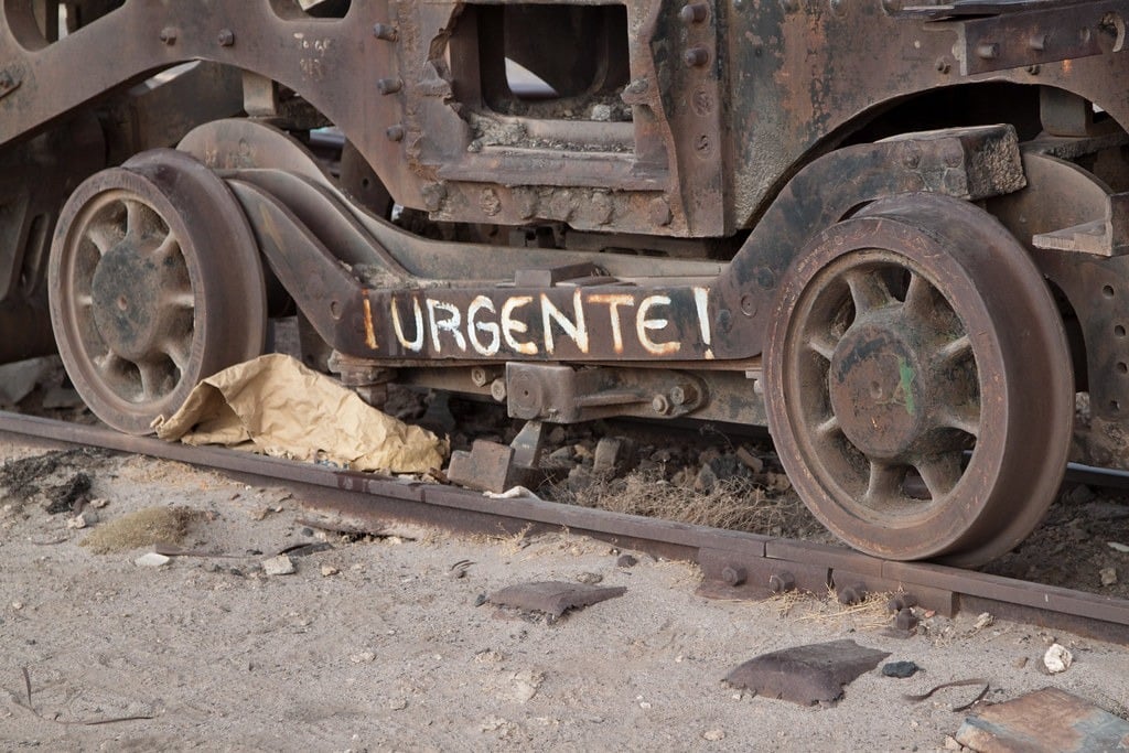 Closeup on the wheels of a train car, where graffiti reads, "URGENTE!"