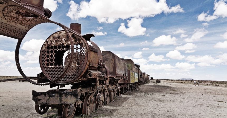 A line of abandoned train cars against a blue sky with clouds in the desert.