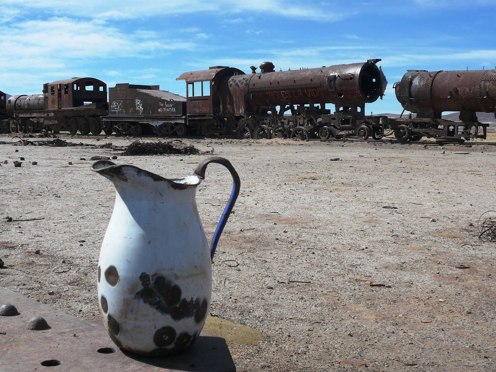 In the foreground, a battered, white clay water pitcher. In the background, abandoned train cars against a blue sky in the desert.