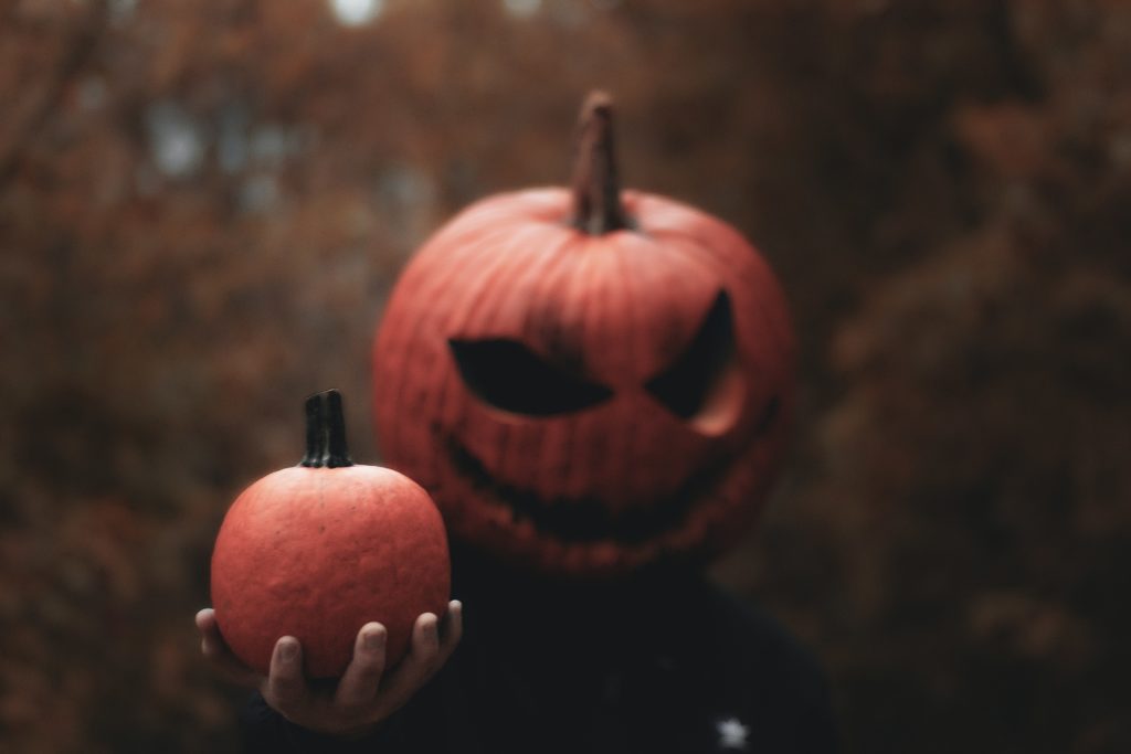 A person with a jack o lantern for a head offering a small pumpkin to the viewer 
