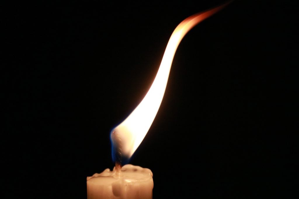 A burning, white candle with tall flame against a dark, black background