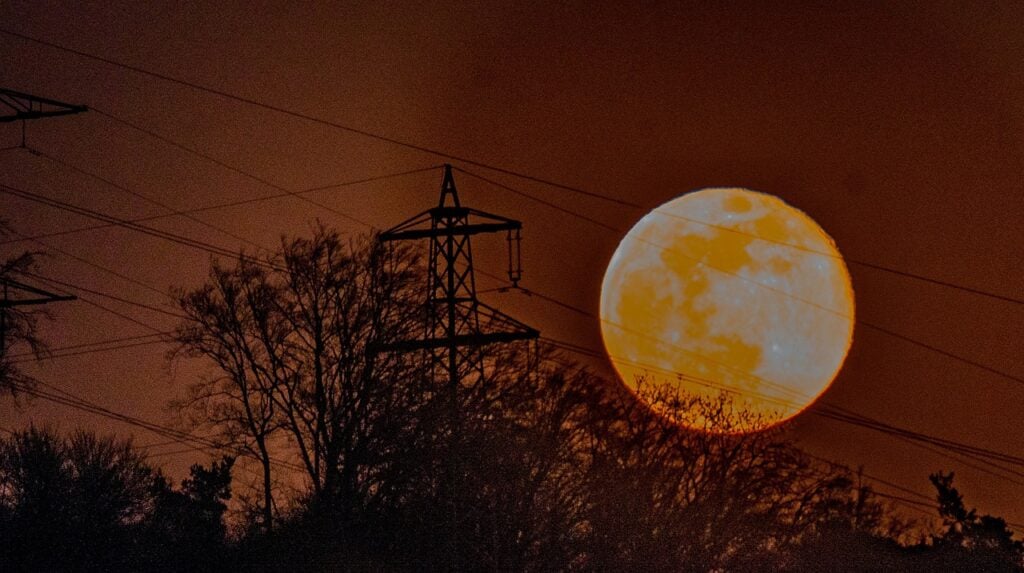 A large full moon against a red sky, with powerlines and trees silhouetted against the light