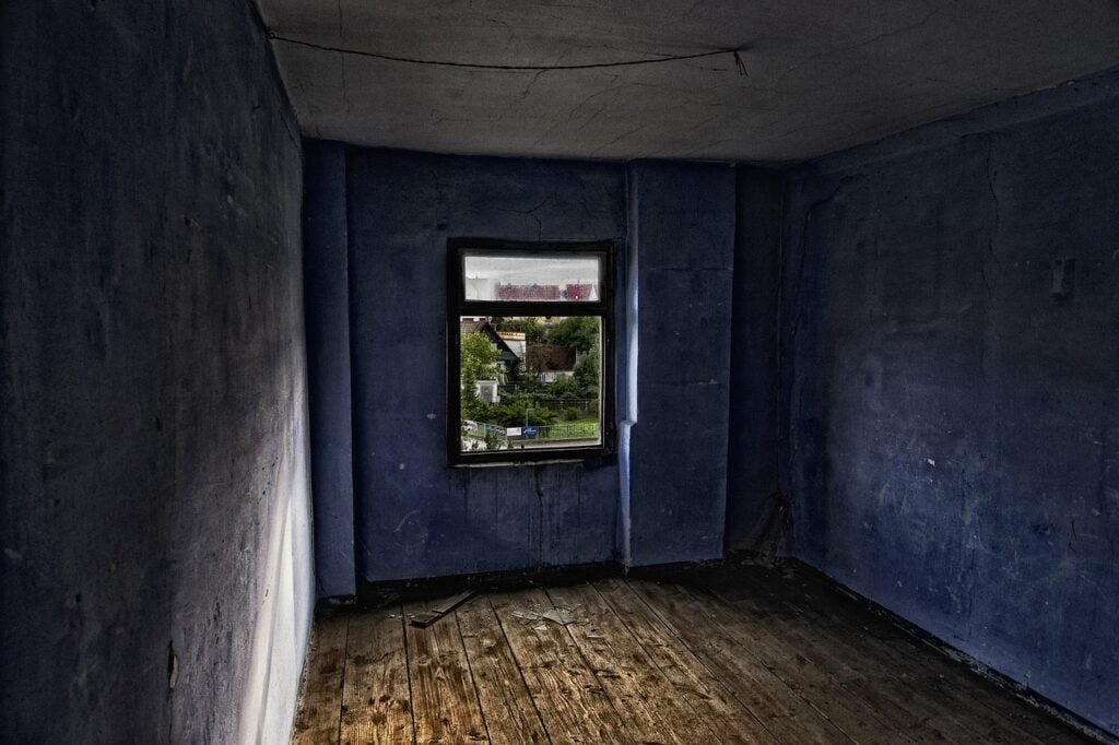 An empty room in an abandoned home with a wood floor, blue walls, and one window