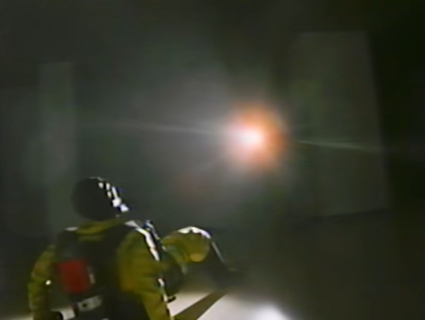 a man in yellow PPE on the floor, looking up at a person pointing a flashlight and weapon at them