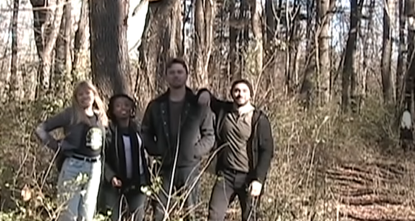 A screenshot from Paranormal Paranoids showing four people - one white woman, one Black woman, and two white men, all in their early 20s - standing in the woods together. There's a ghostly figure to the right in the background.