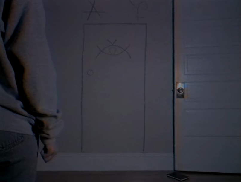 A screenshot from the web series DOORS depicting a white wall with the image of a door drawn on it while a white woman, Sarah, stands in the foreground