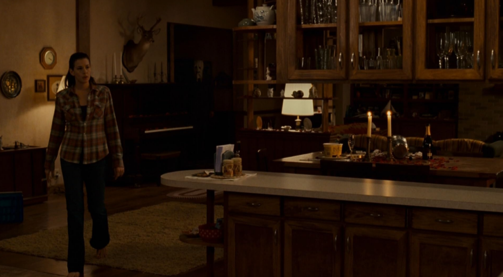 A screenshot from the movie The Strangers depicting Liv Tyler in the foreground inside a cozy cabin and a masked intruder lurking, unseen by her, in the doorframe behind her