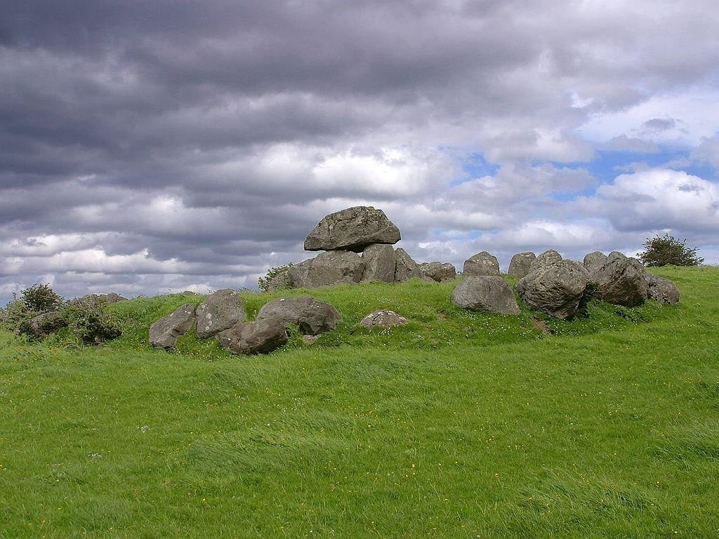 a neolithic tomb or cairn consisting of large stacked rocks in a green field, cloudy sky in the background