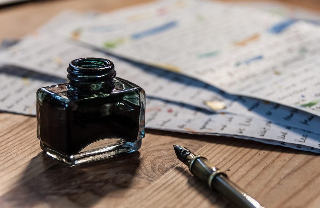 a fountain pen, a bottle of ink, and some scattered papers with writing on them on a wood surface