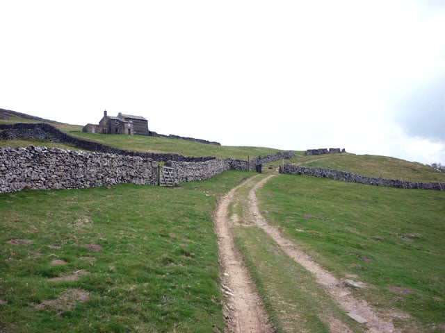 a corpse road, a worn path leading through a green field with a stone wall to the left. an old farmhouse is in the distance