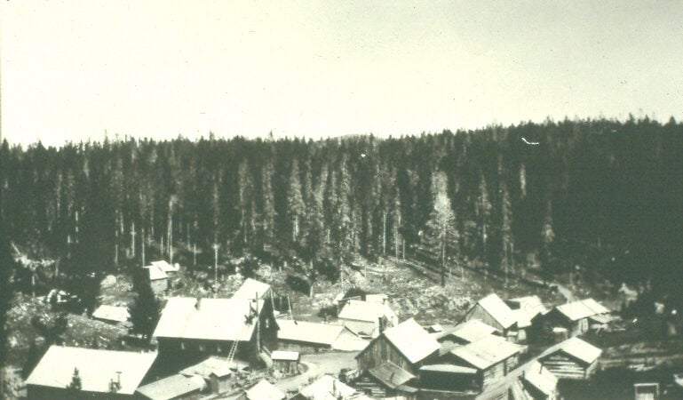 an old photograph of the historic ghost town of Garnet, Montana before it became a ghost town. it consists of several wooden buildings covered in snow and surrounded by pine trees