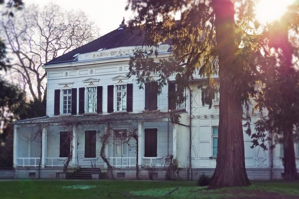 a large, white, two-story house with a porch and a tree out front. it looks abandoned and in poor repair