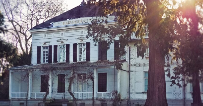 a large, white, two-story house with a porch and a tree out front. it looks abandoned and in poor repair