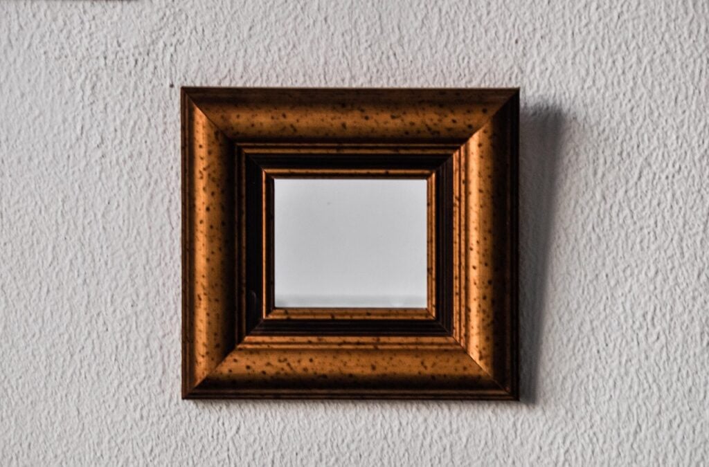 a square mirror with a thick gold frame hanging on a white plaster wall