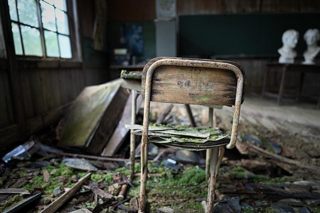 a collapsing desk and chair in an abandoned school. debris and moss all around.