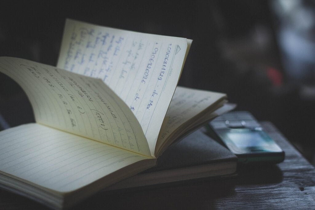an open journal, pages fanned, with writing visible on a table in a dusty, dimly lit room. a cell phone is near by
