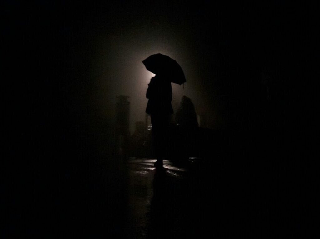 a silhouette of a person holding an open umbrella. the environment is dark, and they are only subtly lit from behind